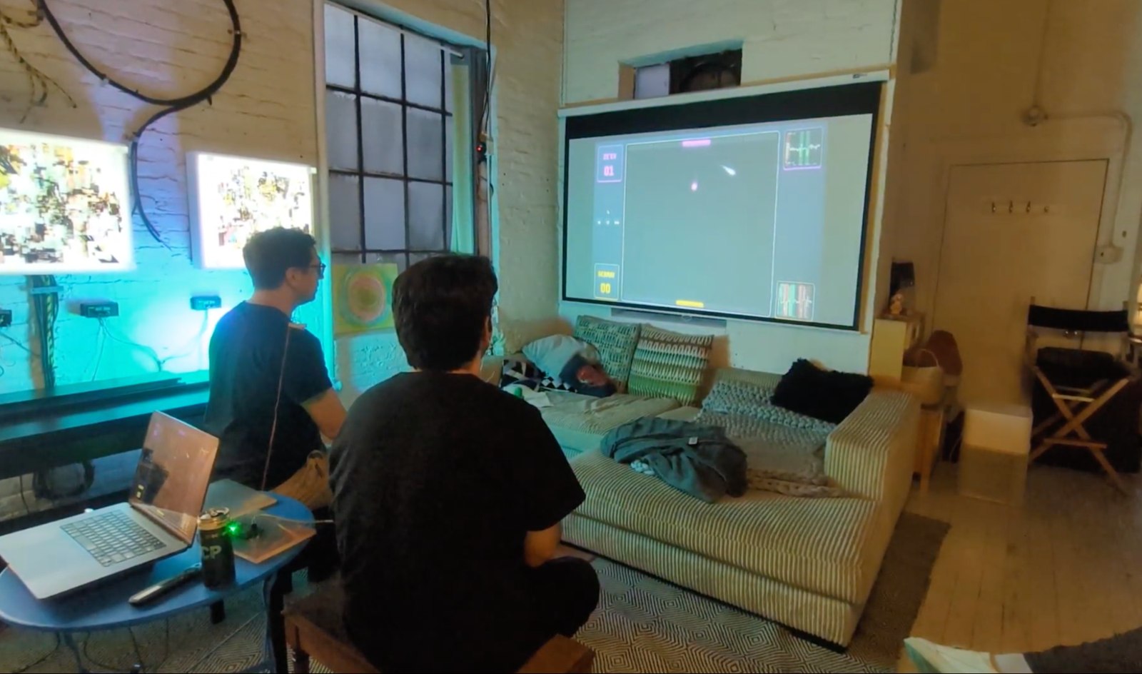 Two competitors seated in front of a projector screen showing the Pong arena mid-game.
